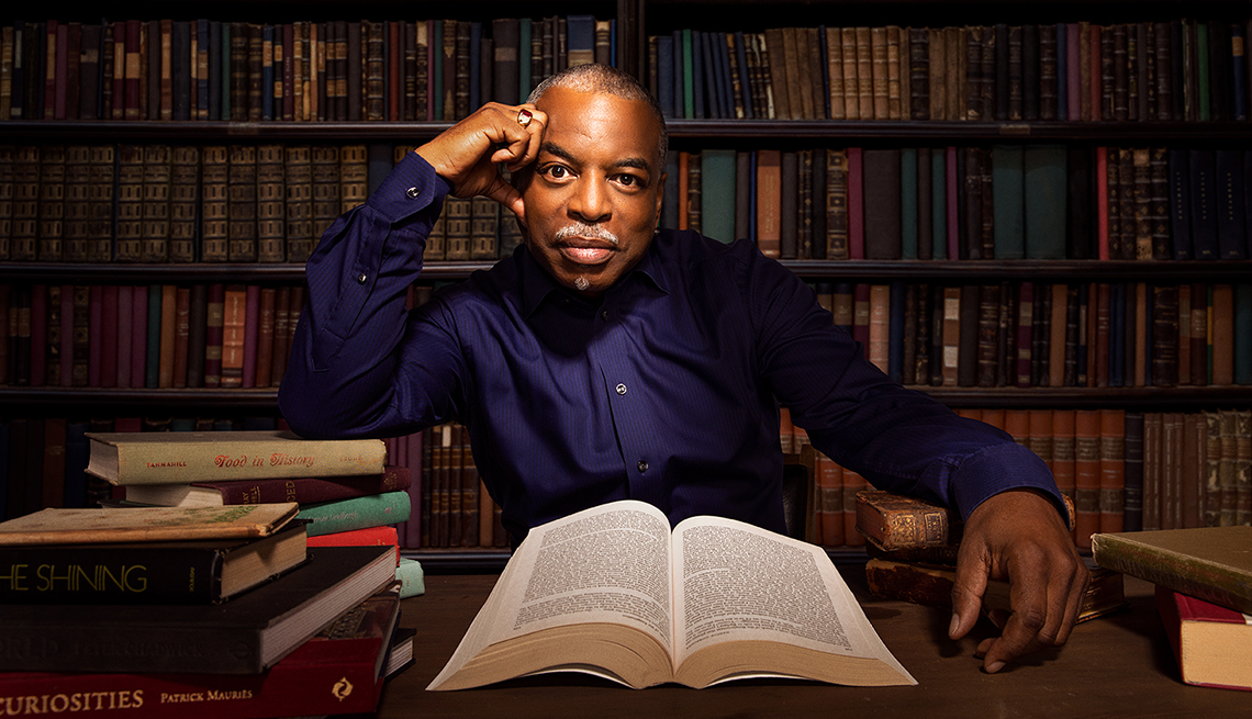 levar burton sitting and leaning head against right hand; book open in front of him; book case with books behind him