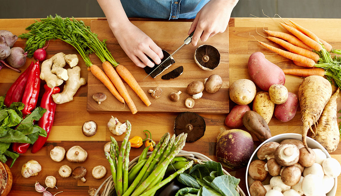 hands chopping a mushroom on a butcher block surrounded by other vegetables including carrots, peppers, potatoes, asparagus and more