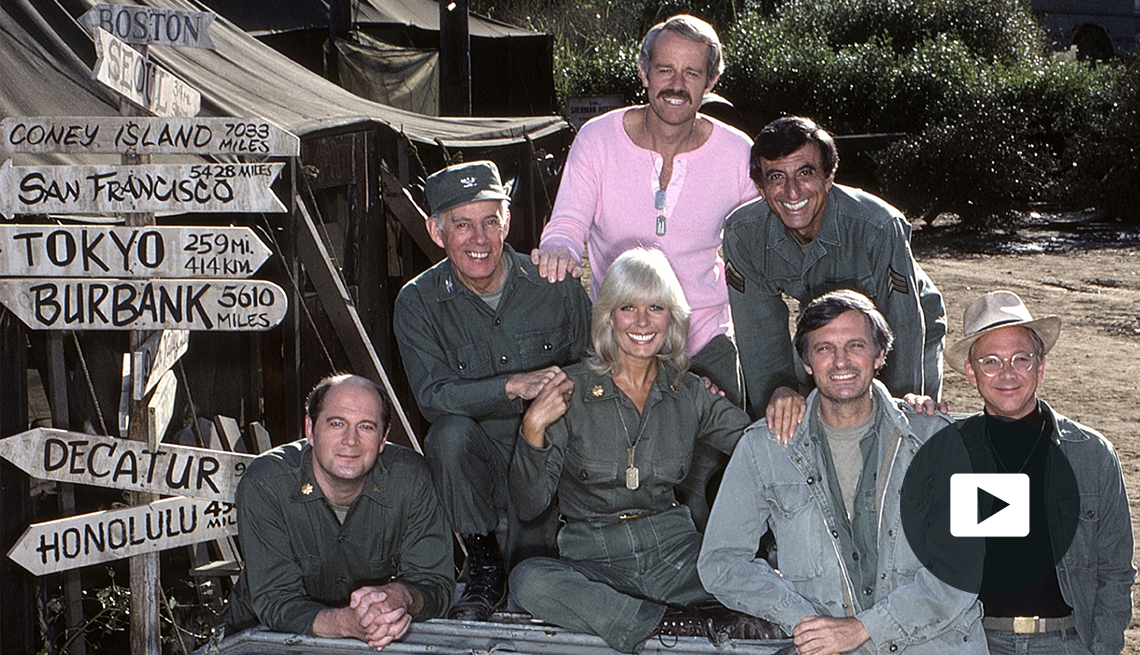M*A*S*H cast members pose for a photo on set next to an army tent and wooden sign showing distances to various cities, with video icon overlay