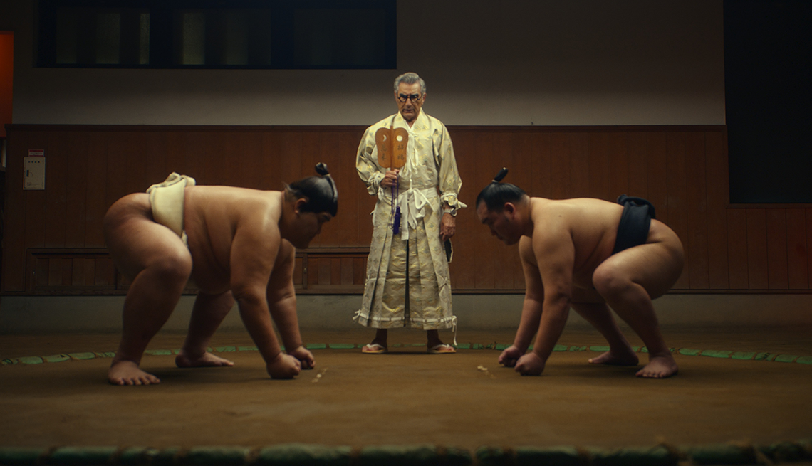 kensho sawada and yoshinori tashiro squatting down in front of eugene levy in a still from the reluctant traveler