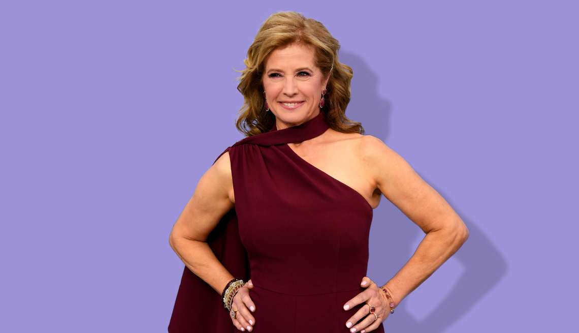 nancy travis with hands on hips in burgundy sleeveless dress against a light purple background