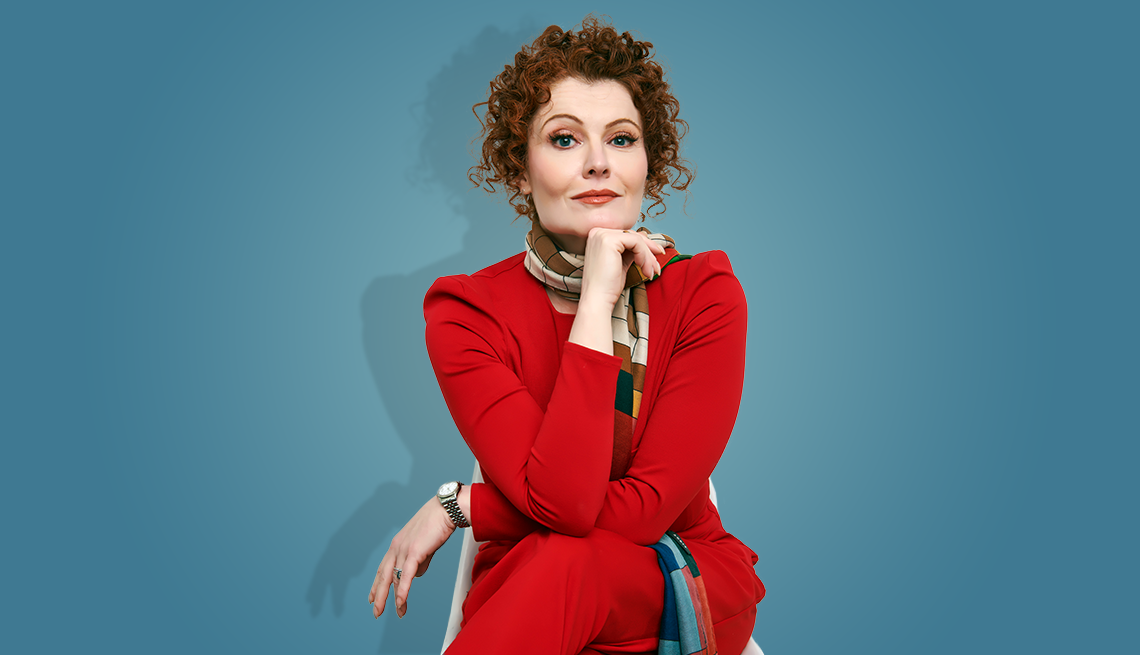 rebecca wisocky in red pants suit with legs crossed and chin resting on right fist; blue background