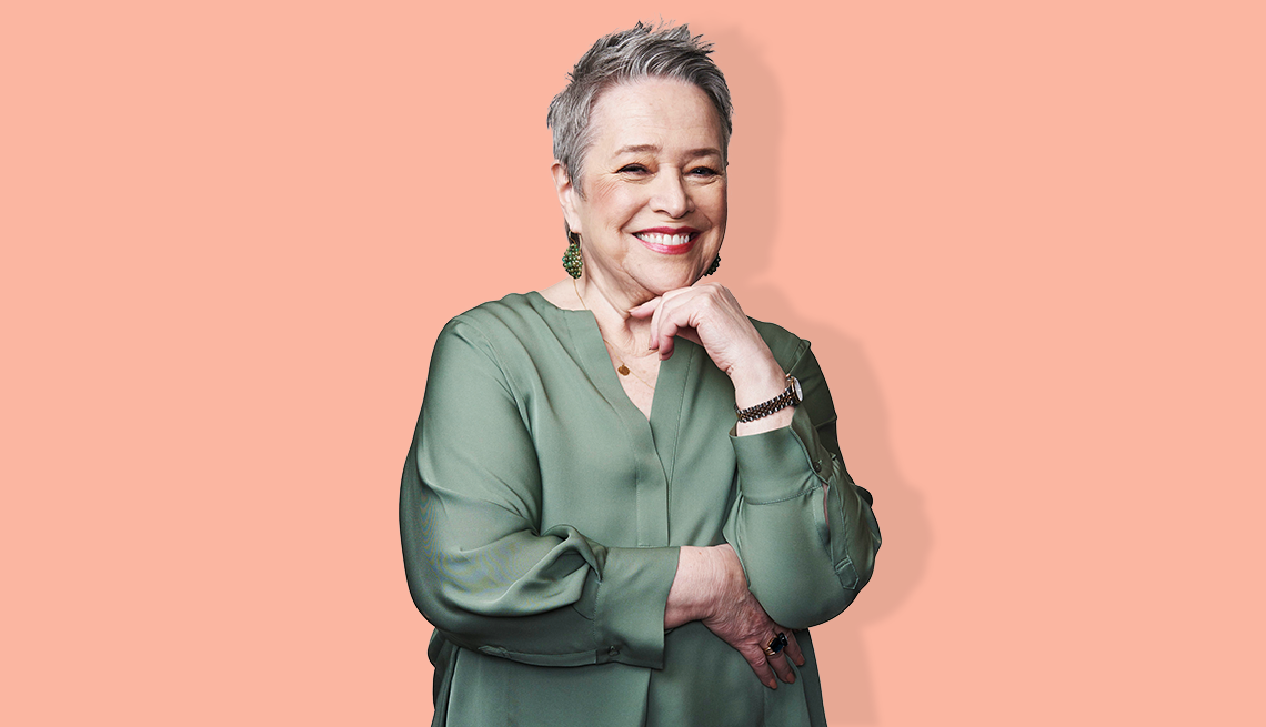 kathy bates smiling with chin resting on her hand against a peach-colored background