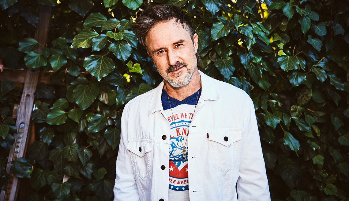 david arquette in a white jean jacket standing in front of a plant-covered wall