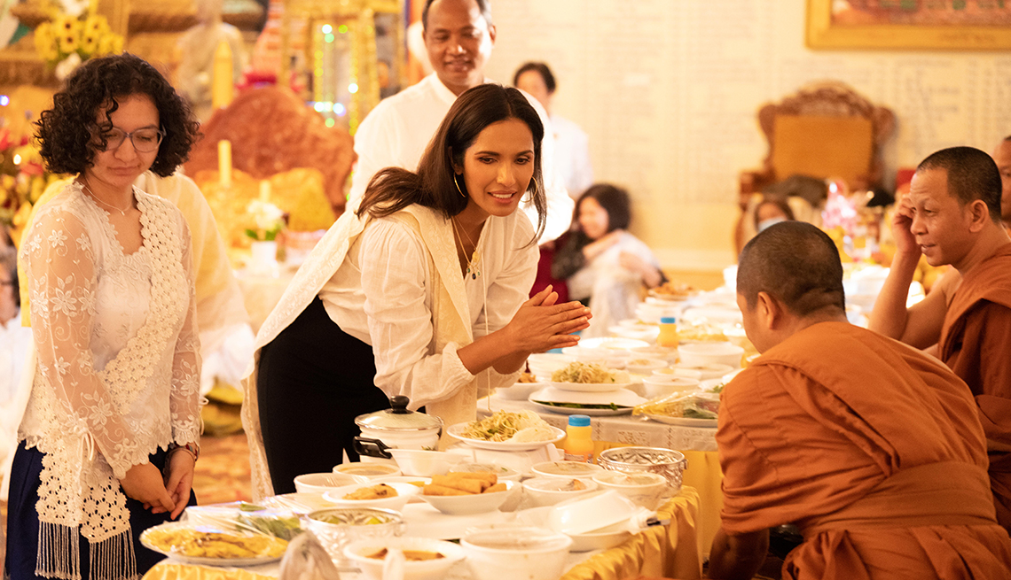 in a still from a taste the nation episode  padma lakshmi with palms together leans over a long table covered with plates of food where two cambodian monks are seated and others are standing or sitting nearby