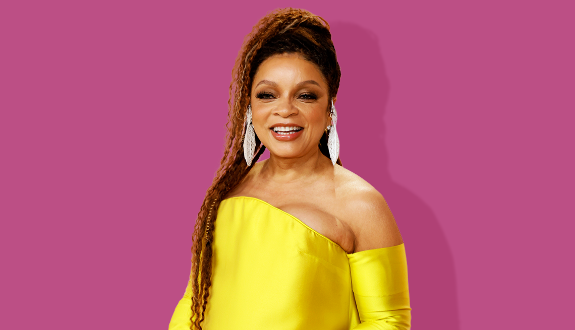 ruth e carter smiling and wearing a yellow dress and long dangling earrings in front of a purple background
