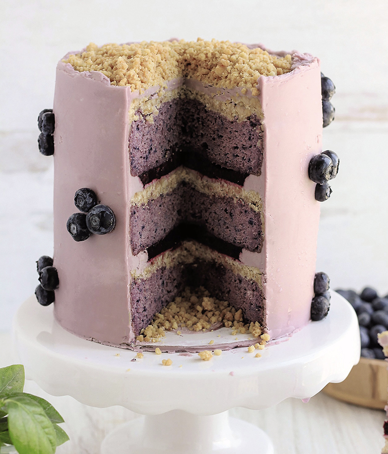 three layer cake with piece cut out; blueberry cake; light pink icing; blueberries on outside of cake; crumble on top