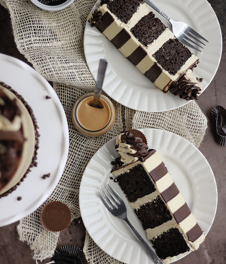 two pieces of three-layer chocolate peanut butter cake on paper plates, each with fork on them; jar of peanut butter with spoon in it is in between the plates; peanut butter cup next to bottom plate