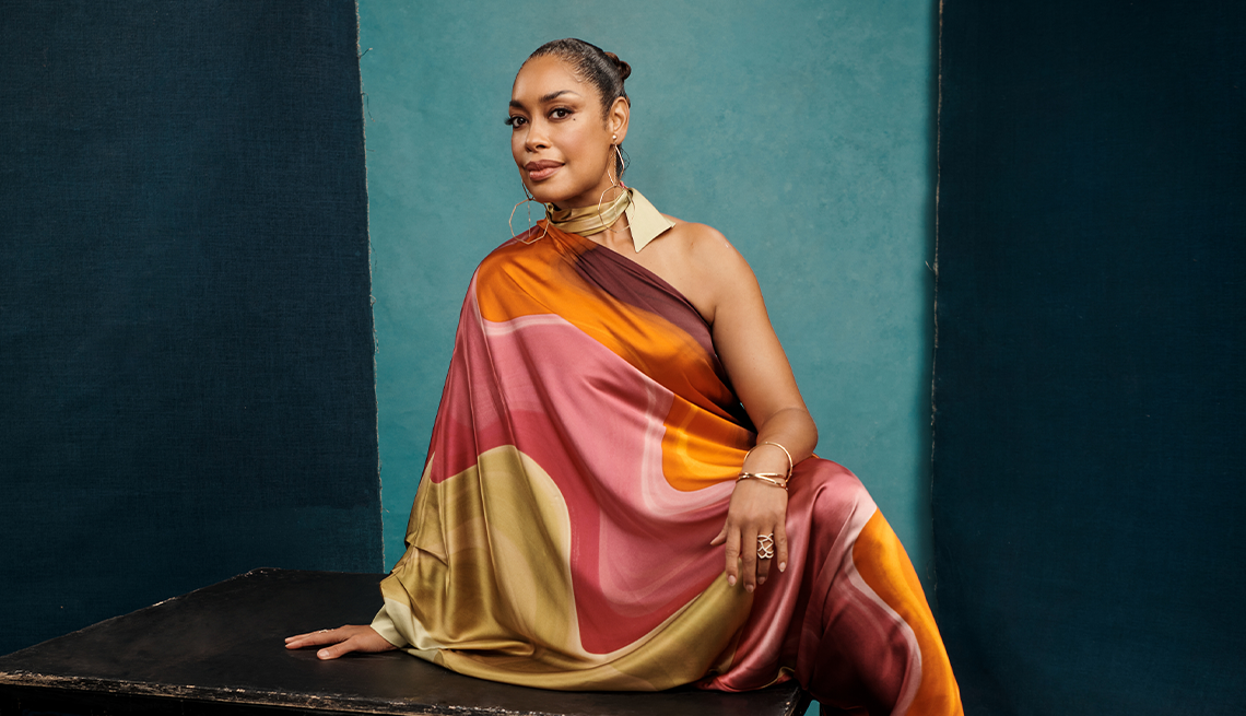 gina torres sits on a black surface in front of a blue and black background, wearing a colorful draping dress and large dangling hoop earrings