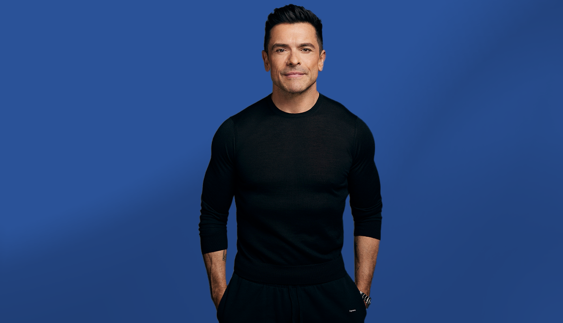 Mark Consuelos standing in all black with hands in pockets; blue background
