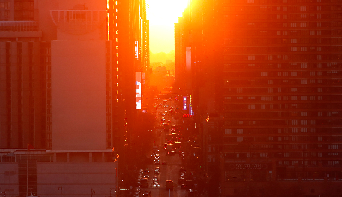 A bright sunrise casts an orange glow between buildings towering over 42nd street in New York City, while cars drive below with their headlights on