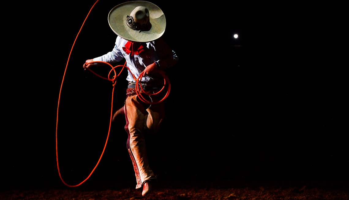 A charro in a broad-brimmed hat with a red handkerchief, a blue shirt and brown leather chaps twirls a bright red lasso against a black field
