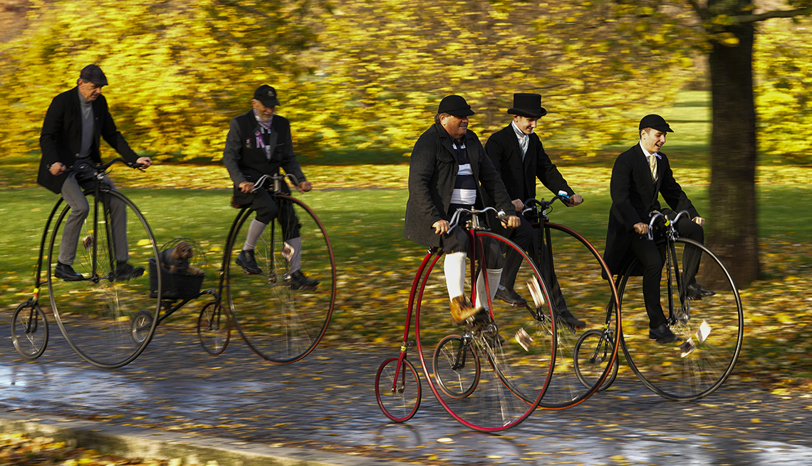 five people dressed in historical costumes ride on their penny-farthing bicycles past vibrant yellow fall foliage