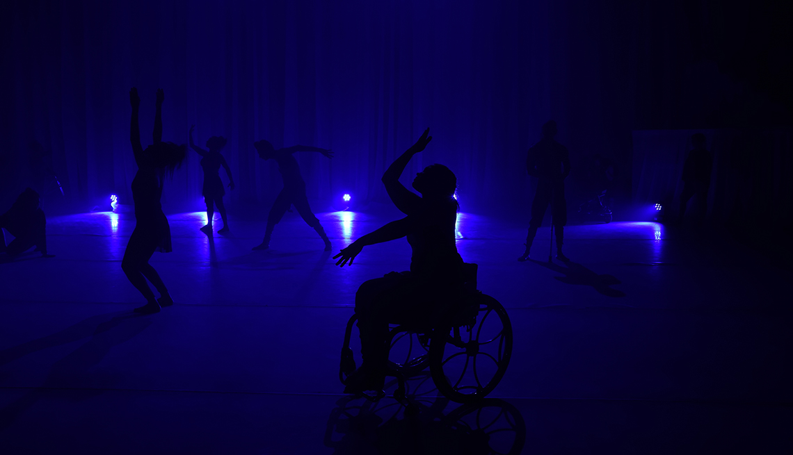 item 15 of Gallery image - silhouettes of a dancer in a wheelchair and other dancers behind on a stage lit with blue light