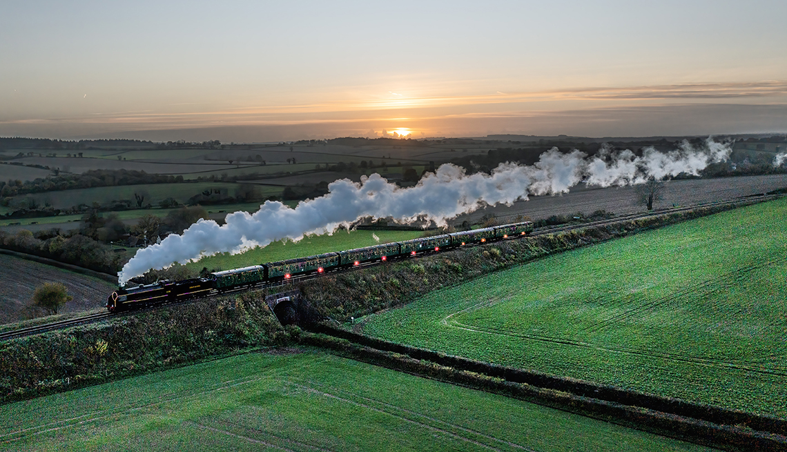 item 12 of Gallery image - a steam train with white smoke billowing behind it passes through green fields with a rising sun in the distance