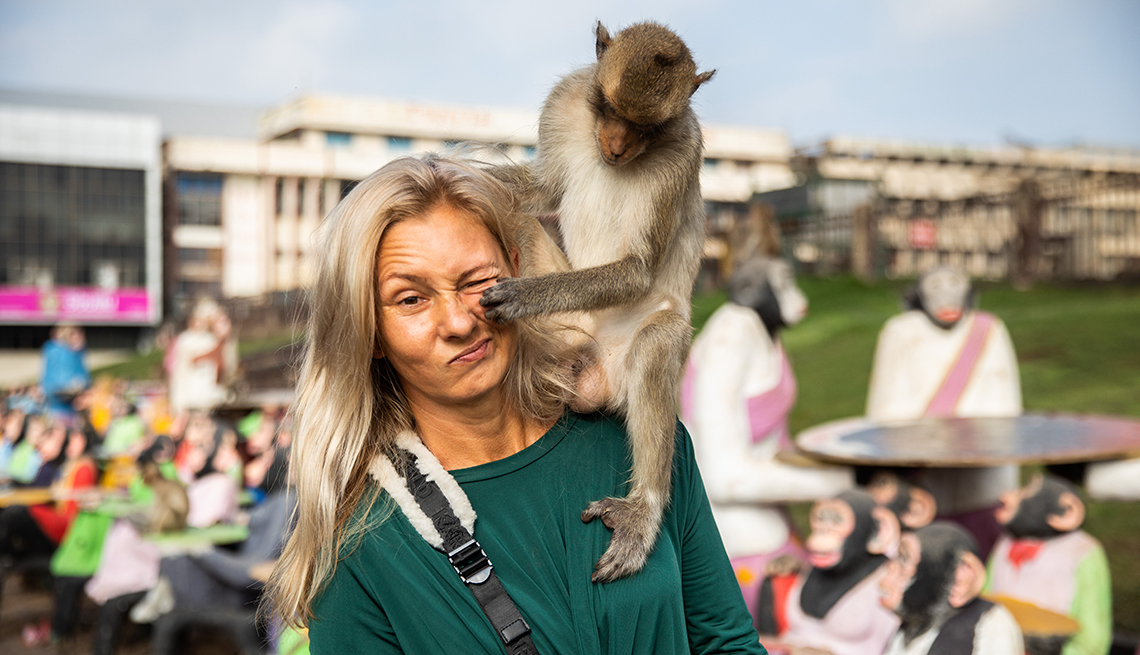 item 6 of Gallery image - a woman makes a funny face as a monkey sitting on her shoulder grabs her cheek, behind them are people wearing monkey masks and buildings in the distance
