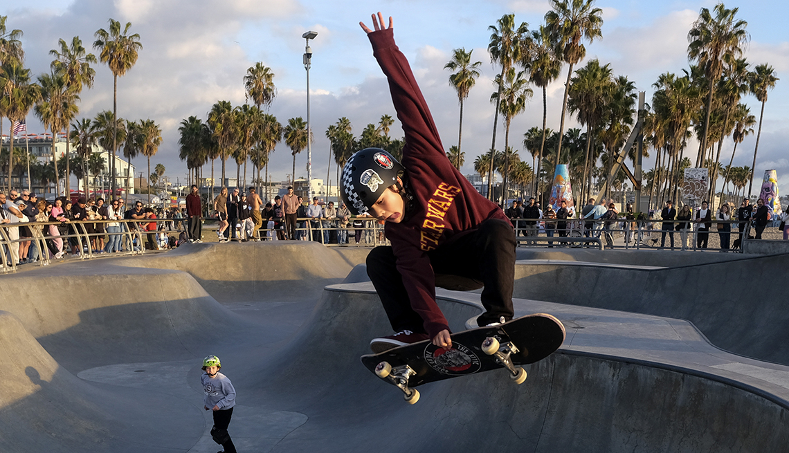 item 9 of Gallery image - a young skateboarder jumps high with his board as a crowd and another skater look on at a skateboard park surrounded by palm tress