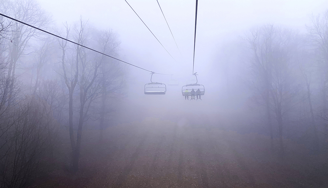 item 12 of Gallery image - three skiers take a chairlift through heavy fog, passing tall trees with bare branches, where no snow is visible