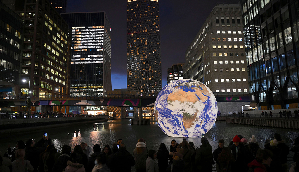 item 11 of Gallery image - people are gathered at london’s canary wharf looking at an art installation that looks like earth floating on the water and glowing against the night sky 