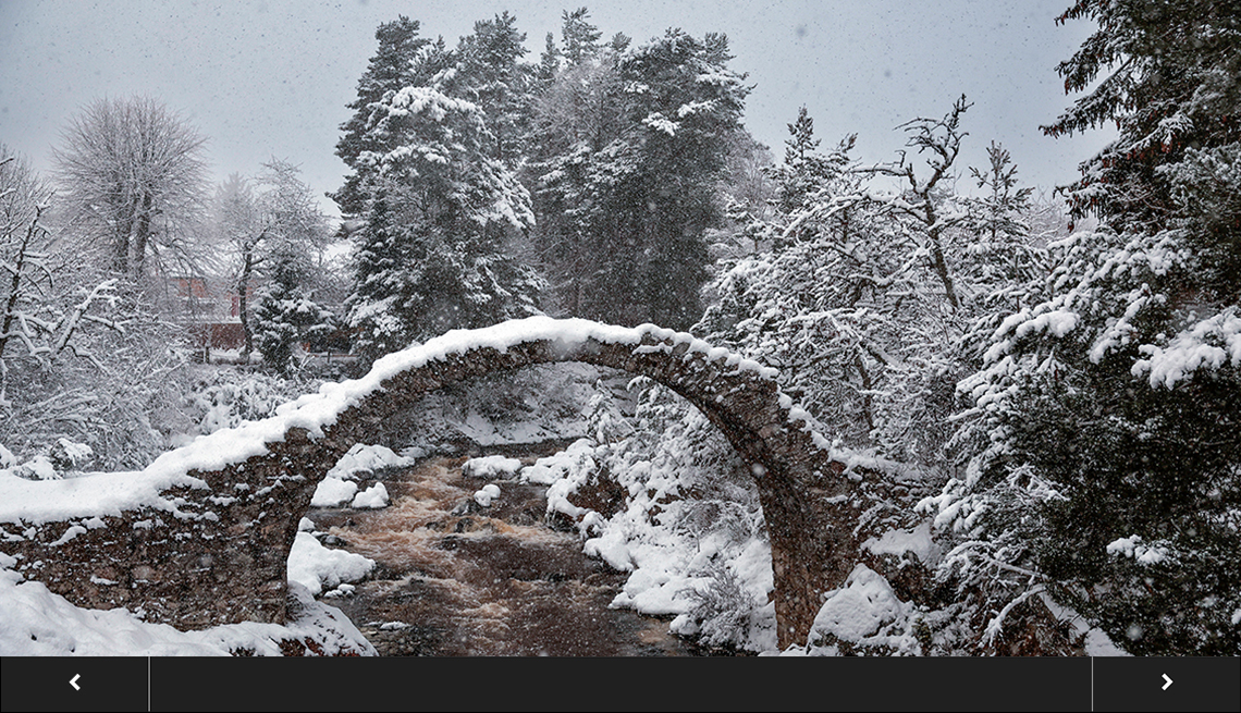 picturesque scene of snow falling on a small stone bridge surrounded by snow-covered trees, with slideshow overlay 