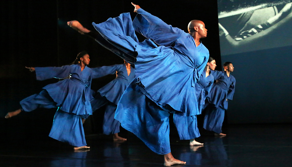 item 7 of Gallery image - five dancers wearing identical flowing blue costumes on a stage, each one in the same pose with one arm and one leg outstretched behind them