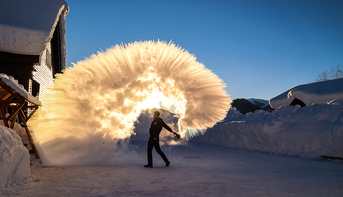 a man standing on a snow-covered street with high snowbanks flings boiling water that turns into ice in midair around him in an arc lit by the sun  