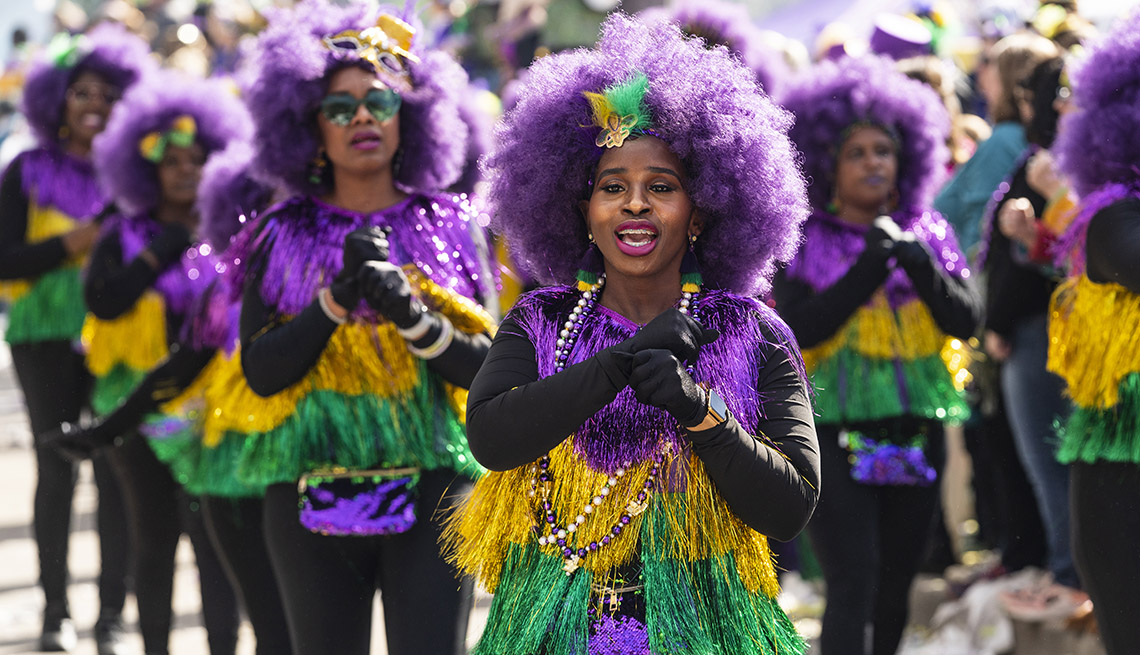 parade participants wearing big purple wigs, beads, and purple, gold and green costumes march down the street 
