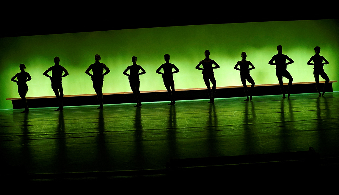 item 17 of Gallery image - nine dancers in identical poses are silhouetted against a backdrop illuminated by a green light