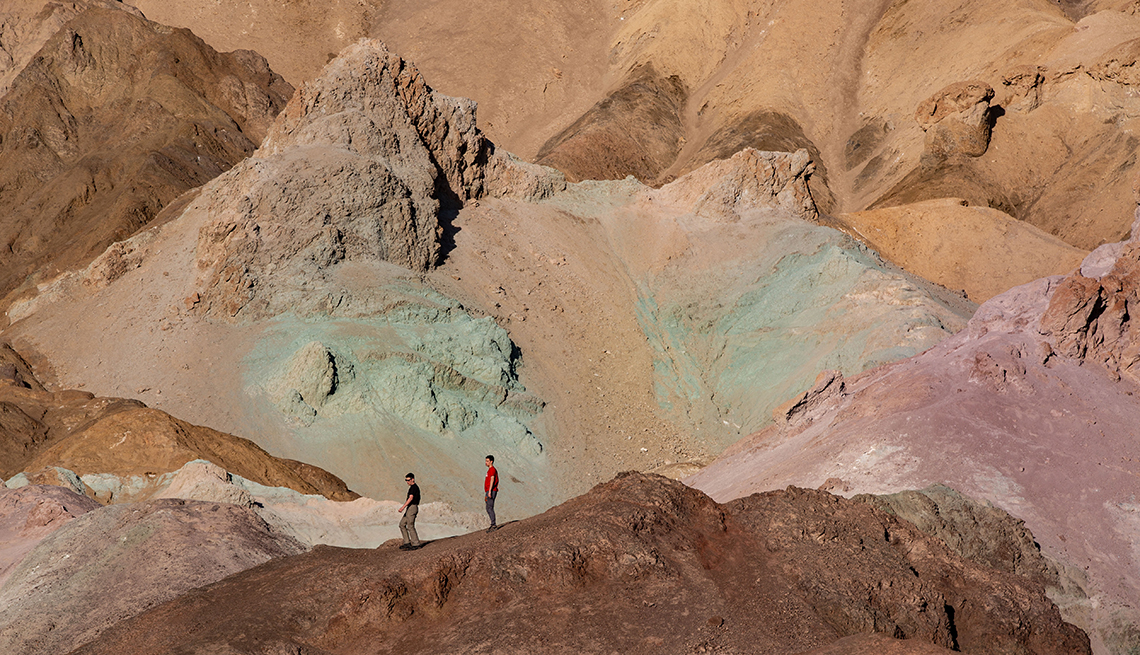 item 11 of Gallery image - two hikers look tiny on a trail with a backdrop of multicolored rock formations of different shades of browns, green and pink