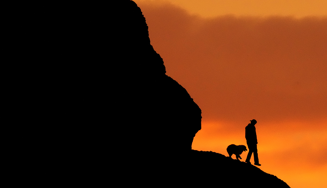item 4 of Gallery image - a man and a dog and the ridge they are hiking along are silhouetted against the orange sunset 