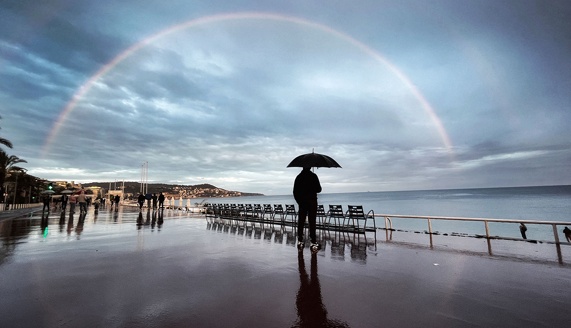 a person wearing black and holding a black umbrella stands on a promenade under a cloudy sky and double rainbow 