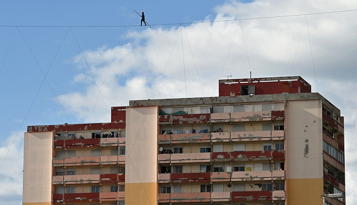 item 4 of Gallery image - a tightrope walker is seen on a highline silhouetted against blue sky and white clouds in front of a shorter apartment building