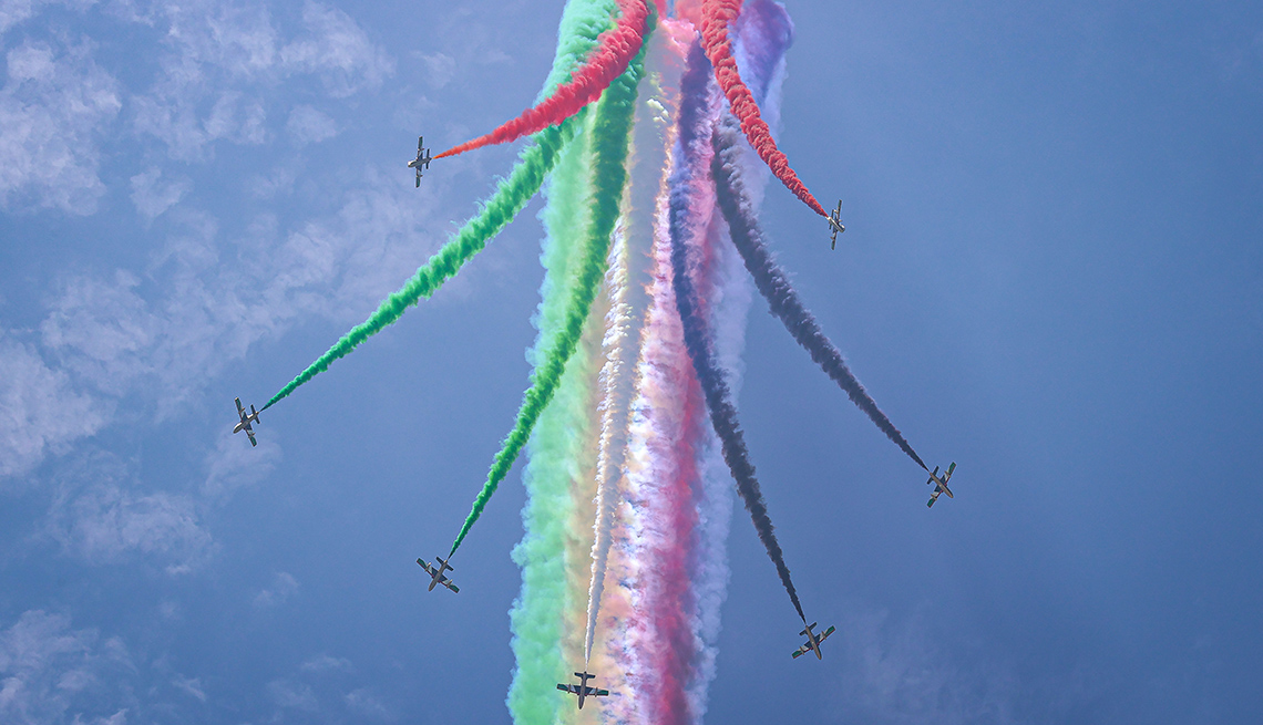 item 15 of Gallery image - seven planes emit trails of red, green, purple and white smoke against blue sky in an aerobatic demonstration 
