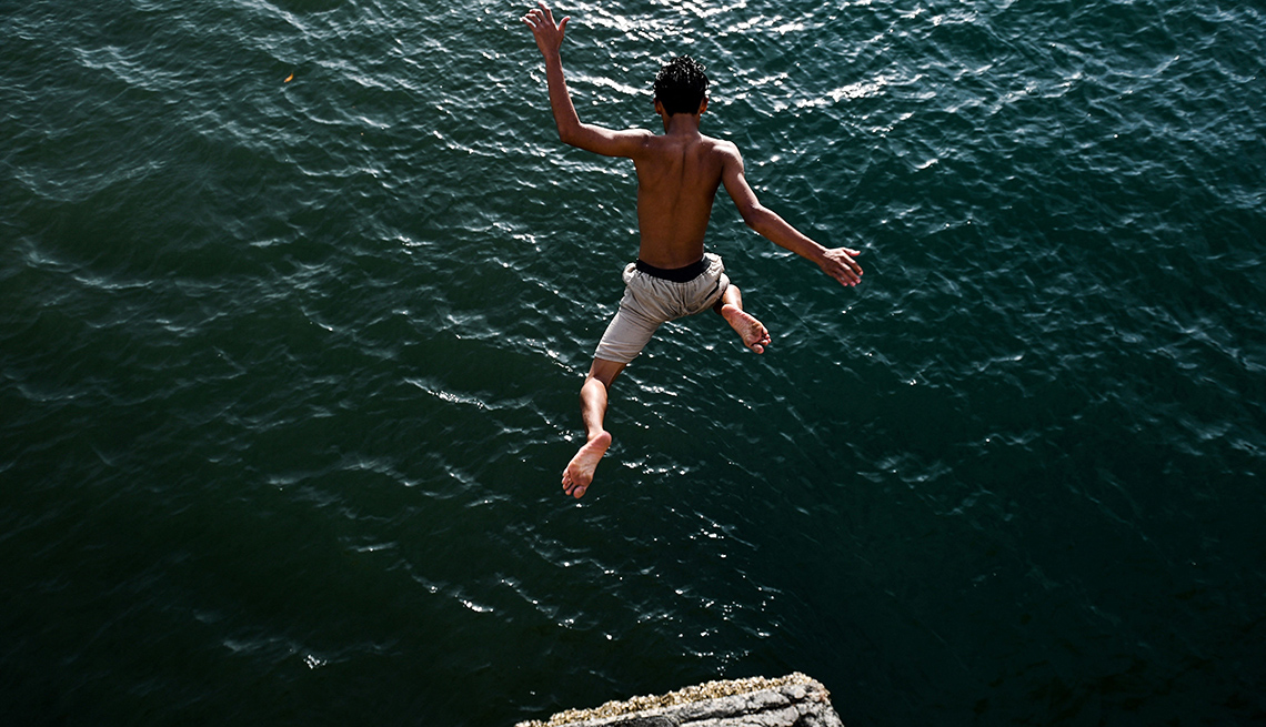 item 4 of Gallery image - a boy jumps into the sea from a rocky ledge, where another person is watching