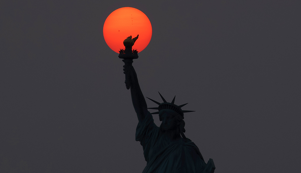 item 12 of Gallery image - the statue of liberty is silhouetted against a hazy sky and the bright orange sun is a perfect circle behind her torch