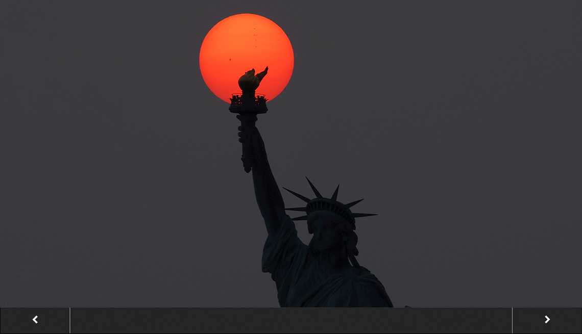 the statue of liberty is silhouetted against a hazy sky and the bright orange sun is a perfect circle behind her torch, with slideshow overlay