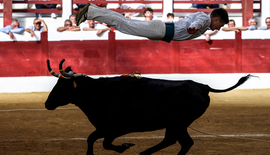 a jumper dressed in white is suspended parallel to a bull running in the opposite direction underneath him as spectators watch in a bullring 