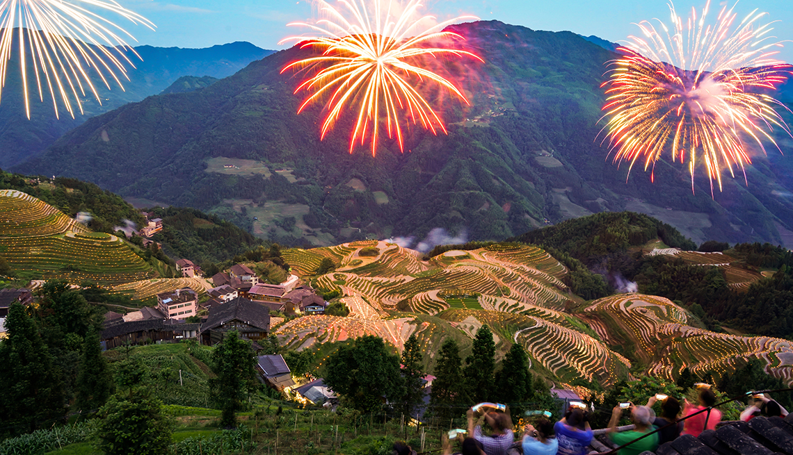 people are seen on a balcony watching fireworks explode over mountain-backed rice terraces and houses