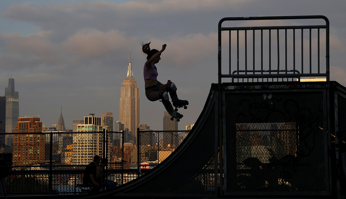 The sun sets on midtown Manhattan and the Empire State Building in New York City as a person does a trick on roller skates at a park in Hoboken, New Jersey. 