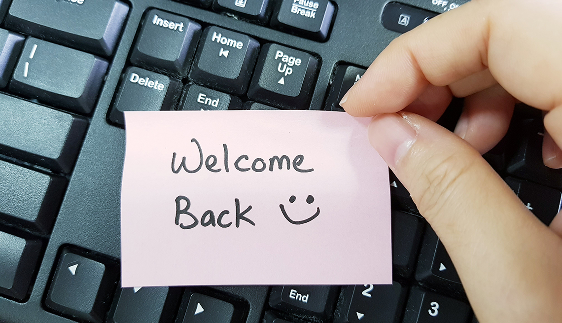 Fingers hold a pink sticky note that says, "Welcome Back" with a smiley face, over a computer keyboard