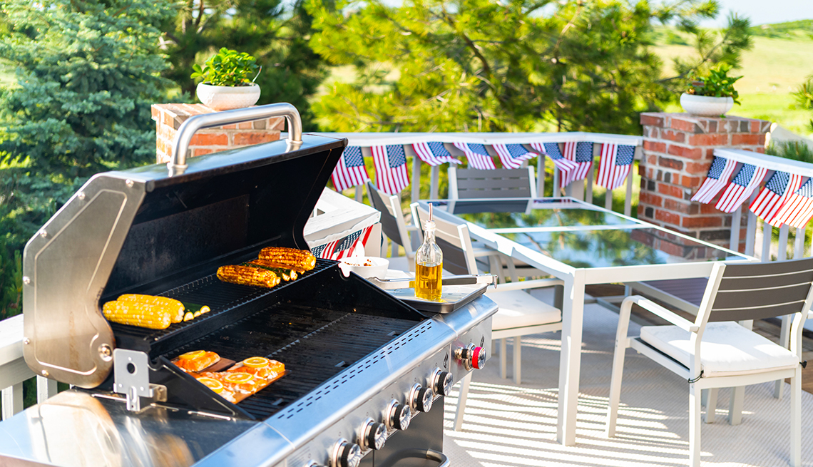 Food grilling on a stainless steel grill. A patio table, chairs, and American flag decor is on the deck railing in the background. 