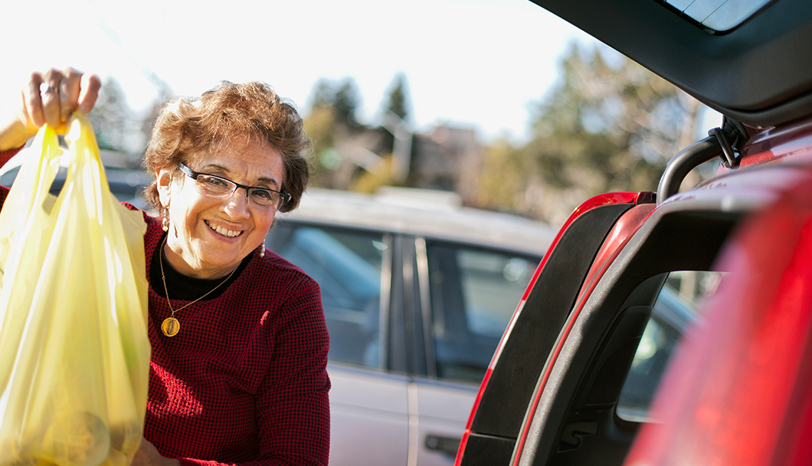 Smiling woman in parking lot holds up bag of groceries next to the opened trunk of her car.
