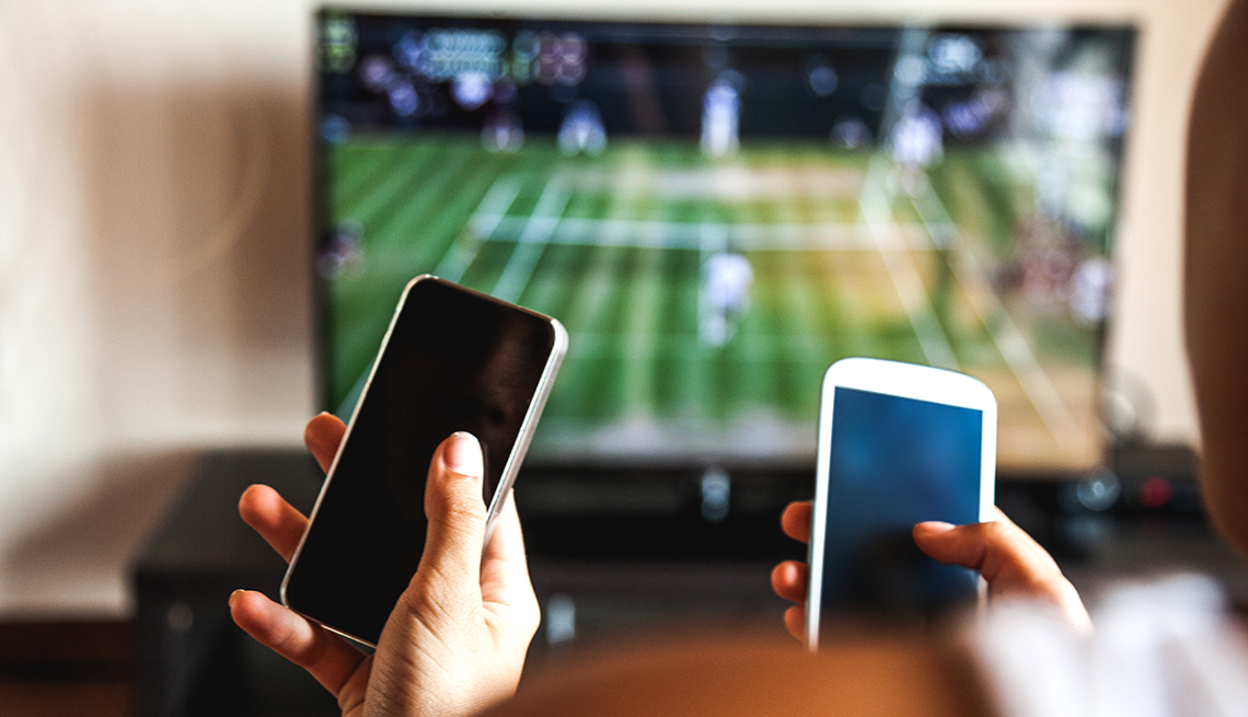 Televisor en la sala de una casa con la transmisión de un partido de fútbol americano y dos manos que sostienen teléfonos móviles