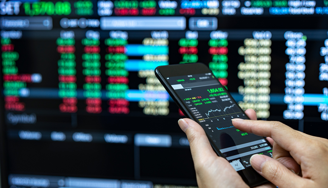 Persona revisando los valores del mercado en un teléfono móvil y al fondo una pantalla con indicadores.
