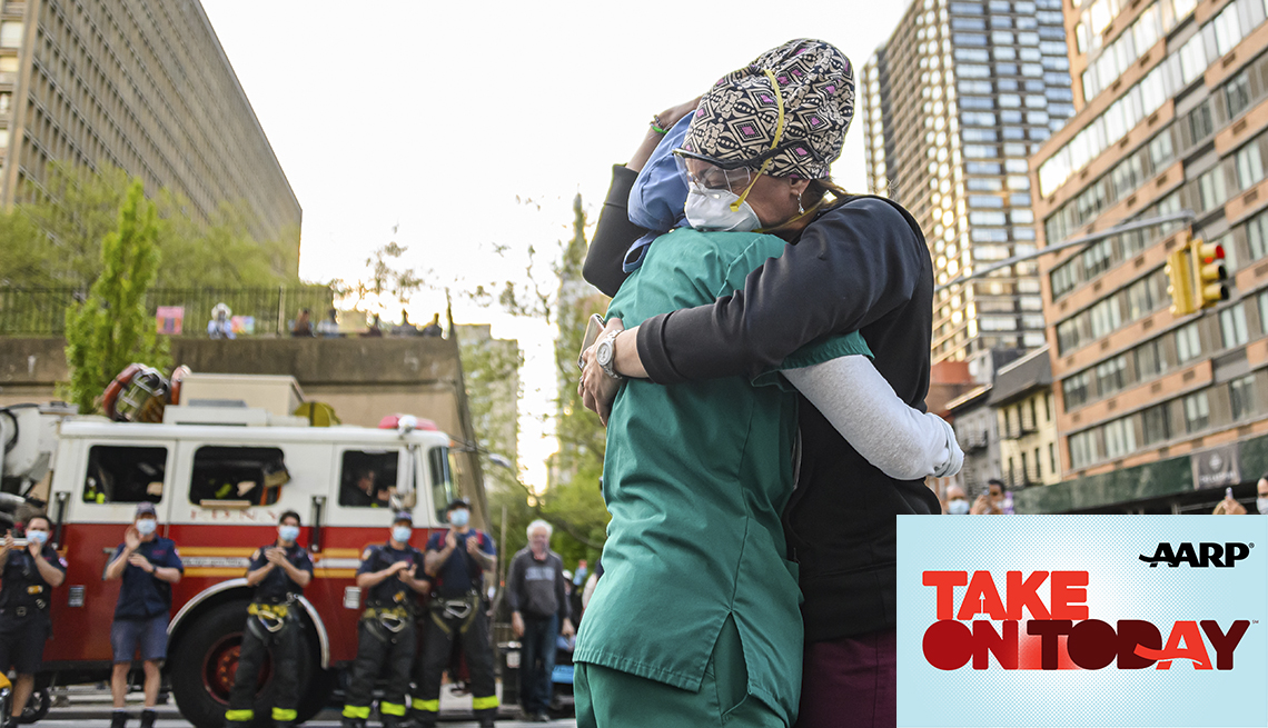 Two nurses hug outside of a hospital with firefighters applauding in the background
