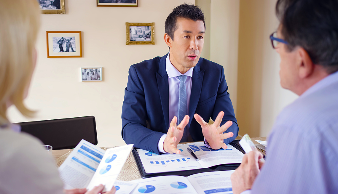 A financial advisor is talking to prospective clients facing him. There are brochures and charts covering the surface of the desk