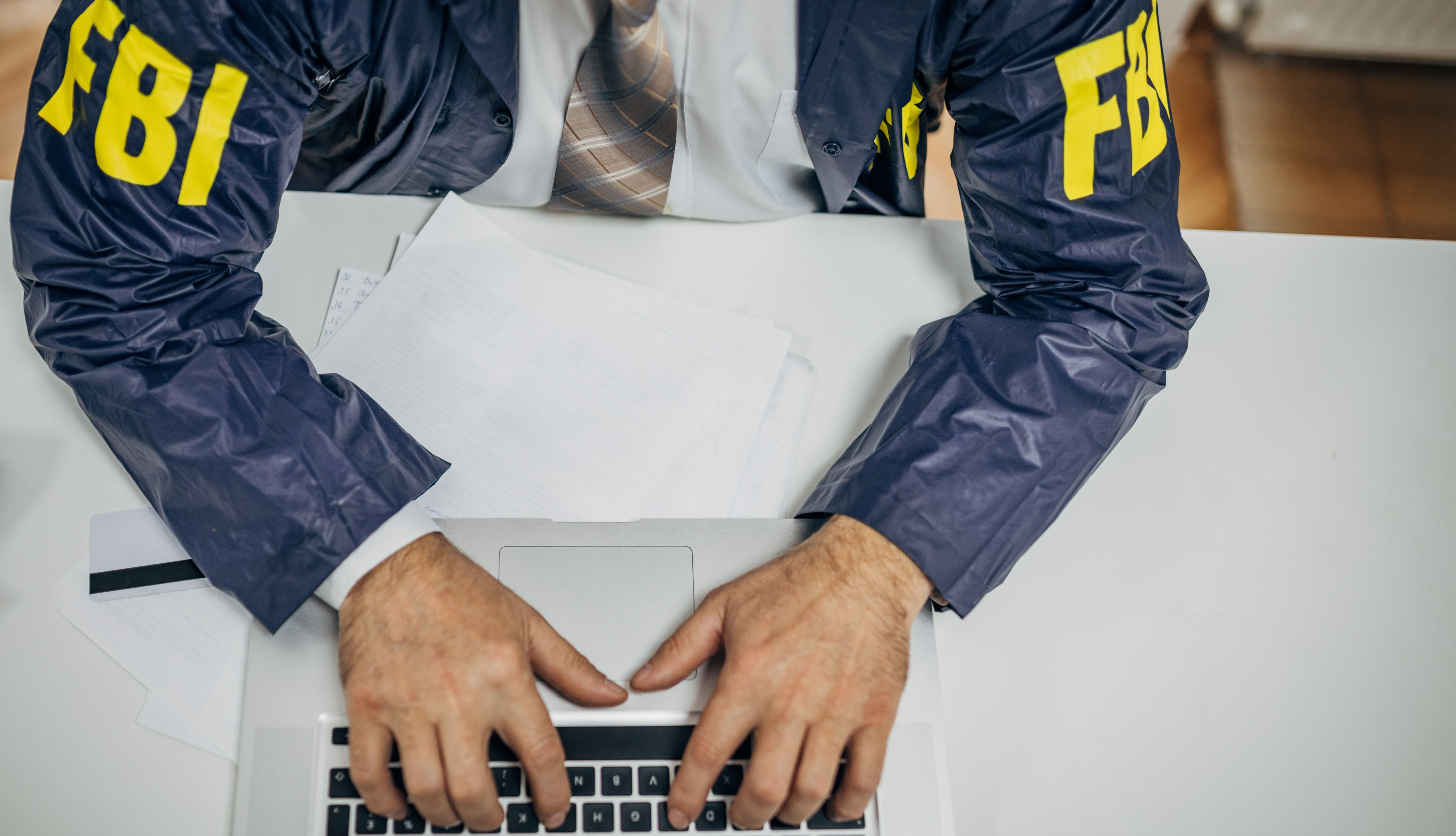 The torso of a FBI agent wearing a blue FBI jacket and typing on his computer.