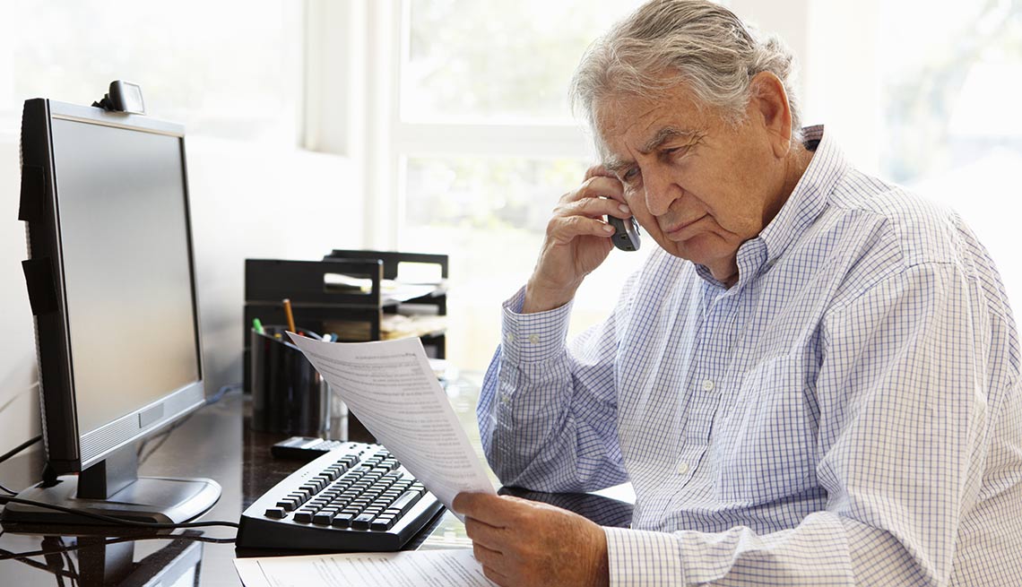 Hombre mayor sentado frente a una computadora sosteniendo un teléfono y unos papeles - Bancos reciben recomendaciones para proteger a personas mayores