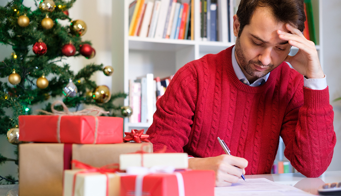 Hombre haciendo cuentas en su escritorio con regalos y un árbol de navidad al fondo.