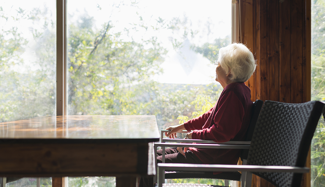 Mujer mayor sentada frente a una mesa mientras mira por la ventana.